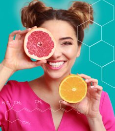 Mulher sorrindo e segurando duas metades de laranja, uma à frente do olho direito. A imagem reforça a presença das vitaminas a, c, d e e em Spax Hair & Nails.