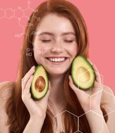 Mulher ruiva com os olhos fechados, sorrindo e segurando duas metades de abacates nas mãos. A imagem reforça a presença das vitaminas do complexo b em Spax Hair & Nails.